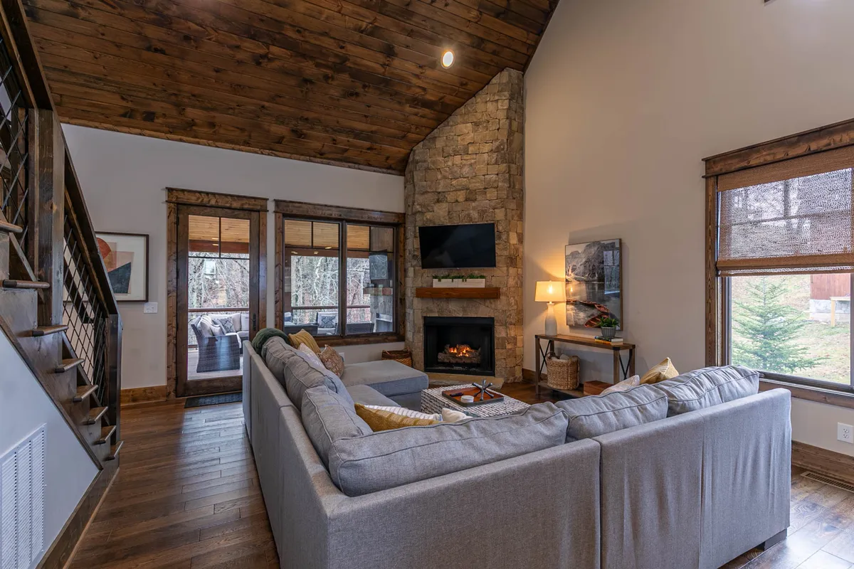 A cozy interior scene with a stone fireplace, large sectional sofa, and big windows allowing natural light to fill the room. The space invites relaxation by the fire, socializing, and enjoying the outdoors from the comfort of inside.