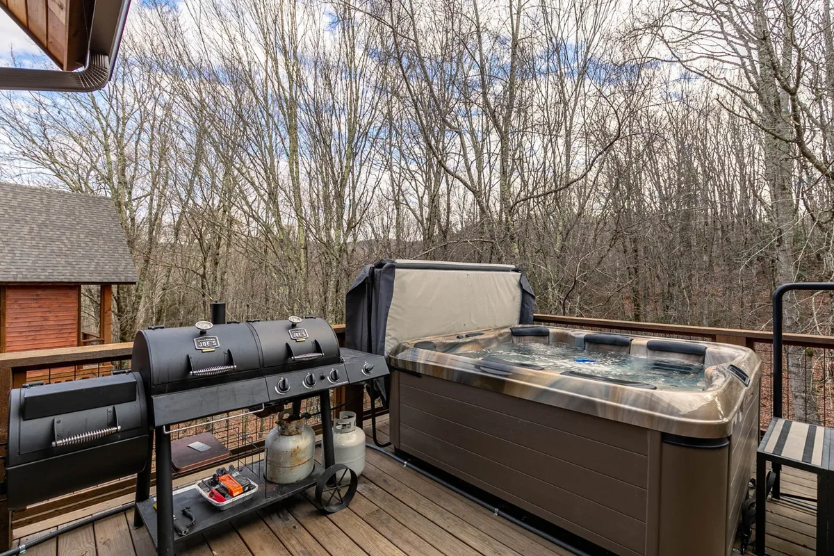 This exterior image features a spacious wooden deck set amidst trees, equipped with a grill and a private hot tub. It radiates a sense of relaxation and leisure, showcasing a tranquil spot for families to unwind after active days, with plenty of space for socializing.