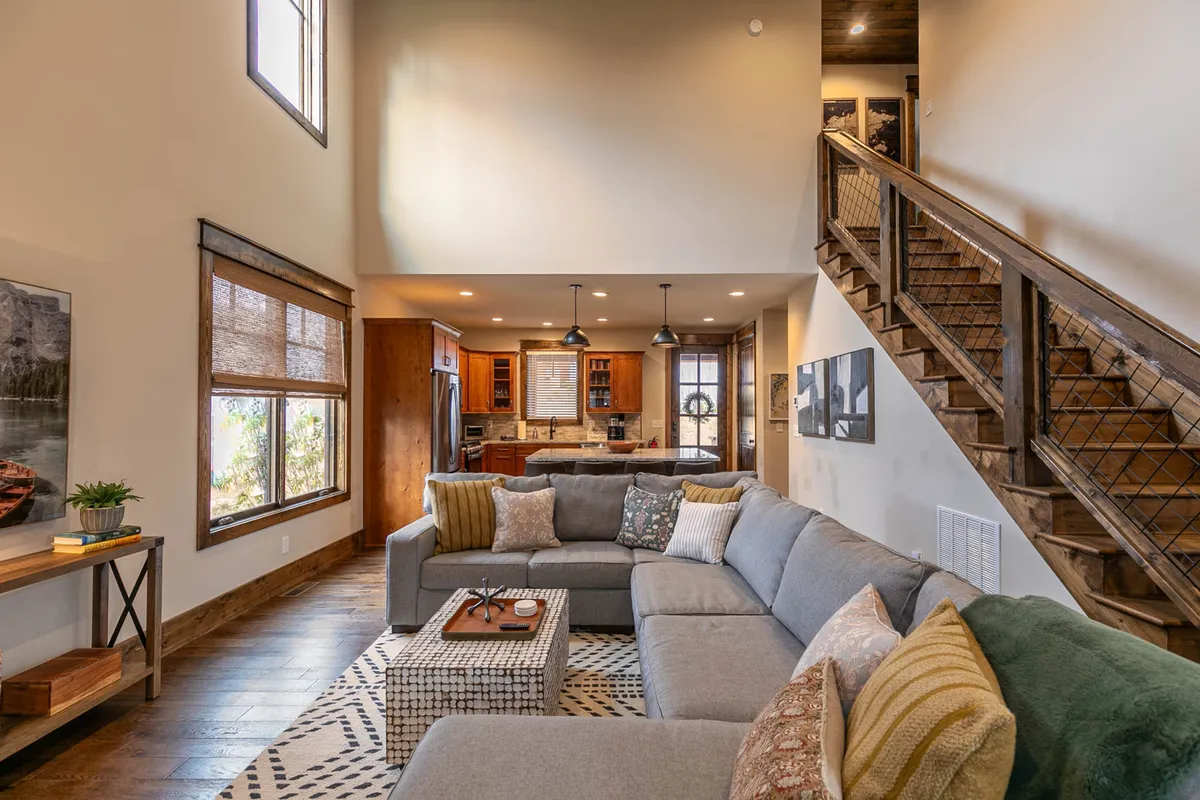 A bright, open-plan living space featuring a large sectional sofa, a coffee table, visible kitchen area, and a stylish staircase. The scene is set up for family movie nights, entertaining, and comfortable relaxation, with natural light streaming in.