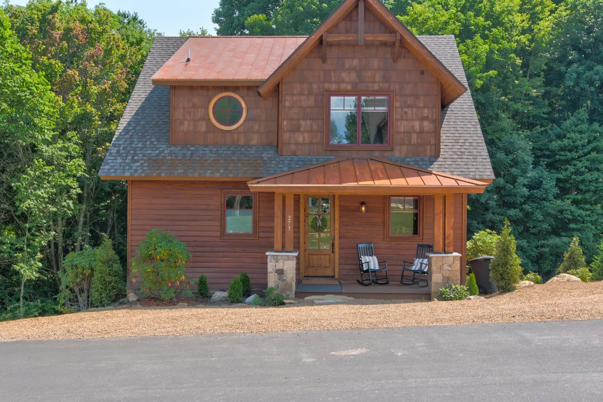 This image depicts the welcoming front entrance of the property, featuring a wooden door, rocking chairs on the porch, and a circular window. The porch is surrounded by natural landscaping, and the design emphasizes a harmonious connection to the outdoors.