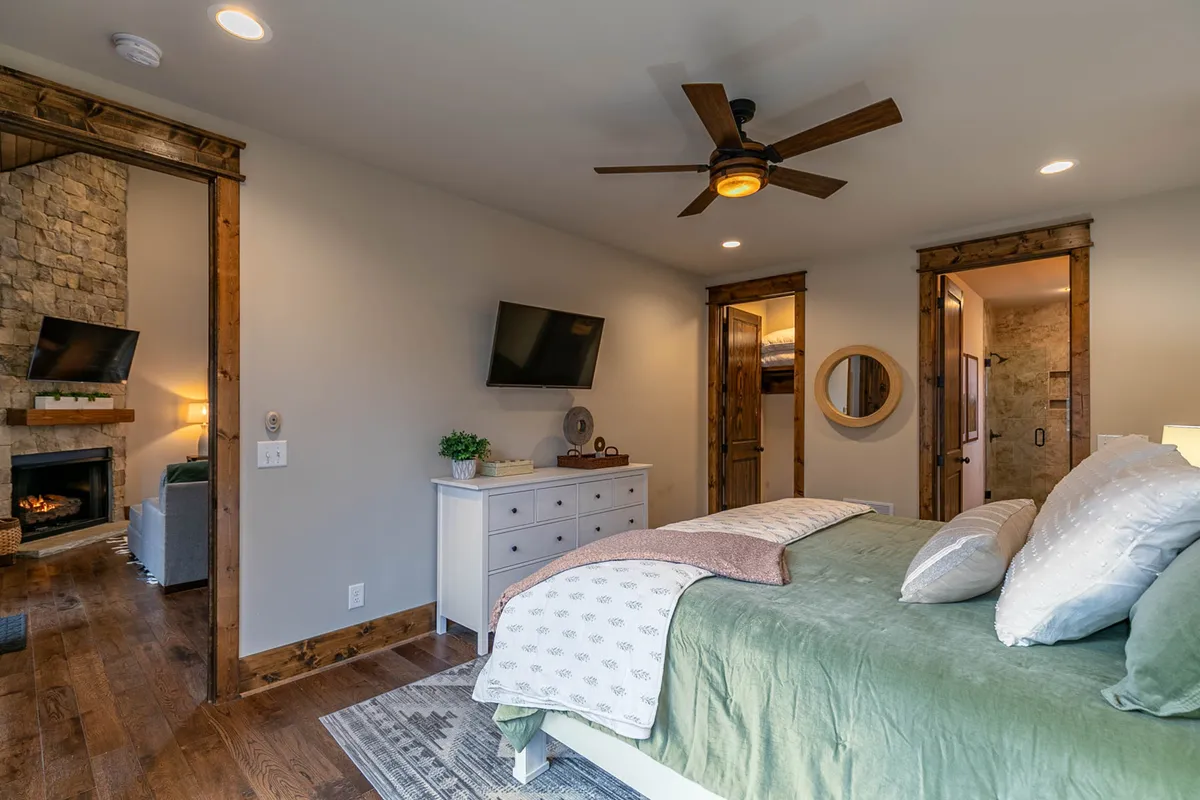 A spacious king bedroom featuring a plush king-size bed draped in luxurious linens, a stone fireplace that adds a cozy ambiance, tasteful wooden furniture, and a wall-mounted TV. Soft natural light filters through the large windows, offering a glimpse of mountain or woodland views and creating a serene atmosphere.
