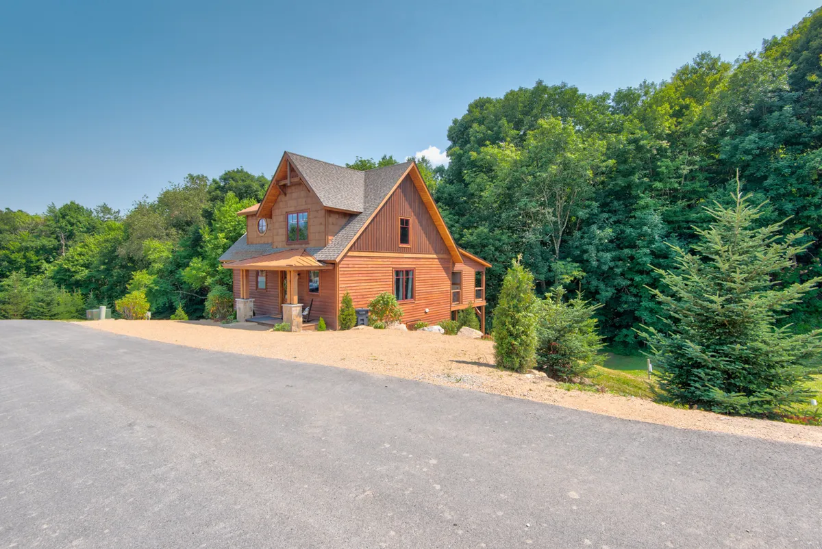 This image features the exterior of a welcoming wooden house surrounded by trees and a gravel driveway, suggesting a spacious setting ideal for gatherings and outdoor activities. The inviting porch hints at a place to relax and enjoy the company of loved ones.