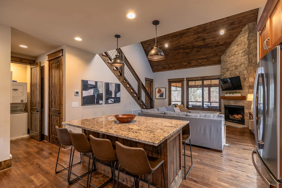 An inviting open-plan interior featuring a spacious granite kitchen island with seating, a modern fireplace, a broad staircase, and expansive windows letting in abundant natural light. The kitchen area boasts gleaming stainless steel appliances, and the living area’s plush seating is oriented for easy socializing and relaxation.