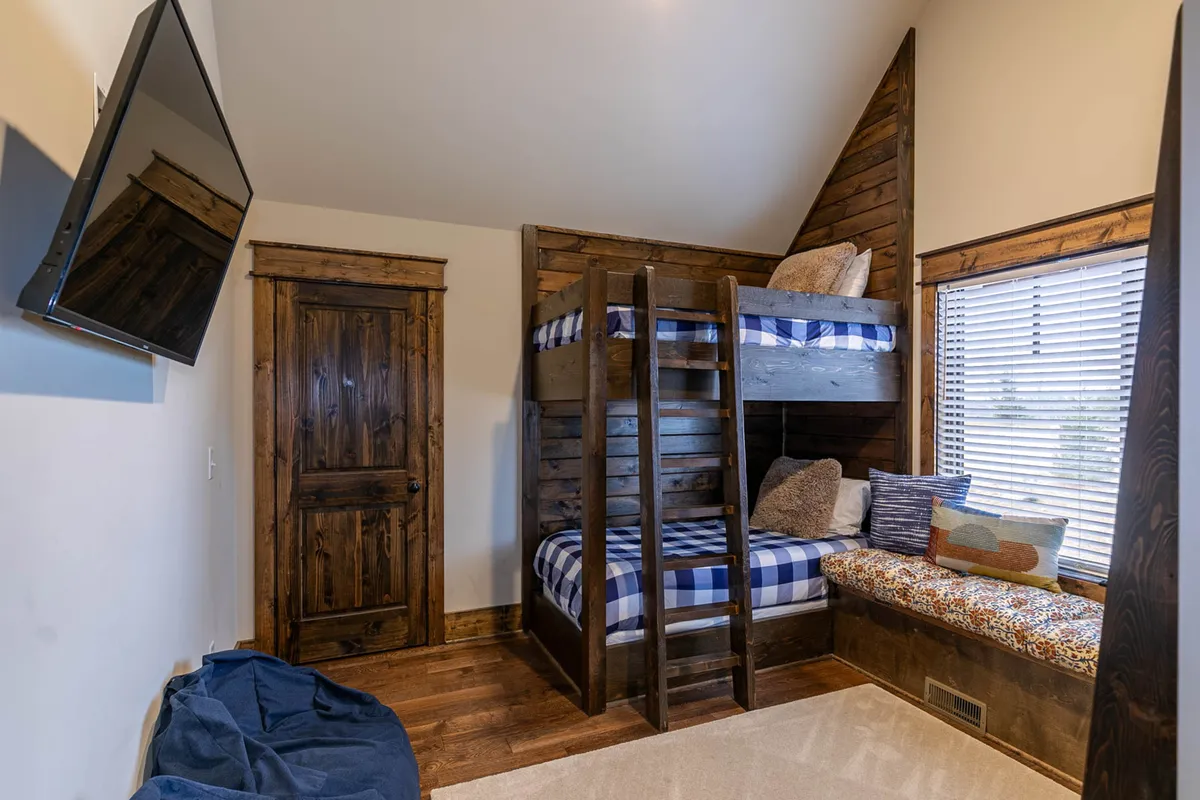 A fun and playful bunk room with two sets of twin bunk beds, a cozy window seat perfect for reading, a TV for movie nights, and a rustic wooden door. Natural light streams in from the window, making the space feel lively and welcoming for kids or groups of friends.