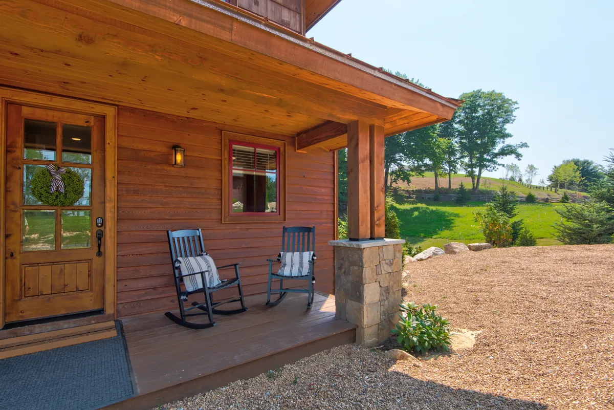 A welcoming front porch features classic rocking chairs, a sturdy wooden door, stone column accents, and lush green landscaping. The scene suggests a quiet morning or peaceful evening spent unwinding while soaking in the mountain air.