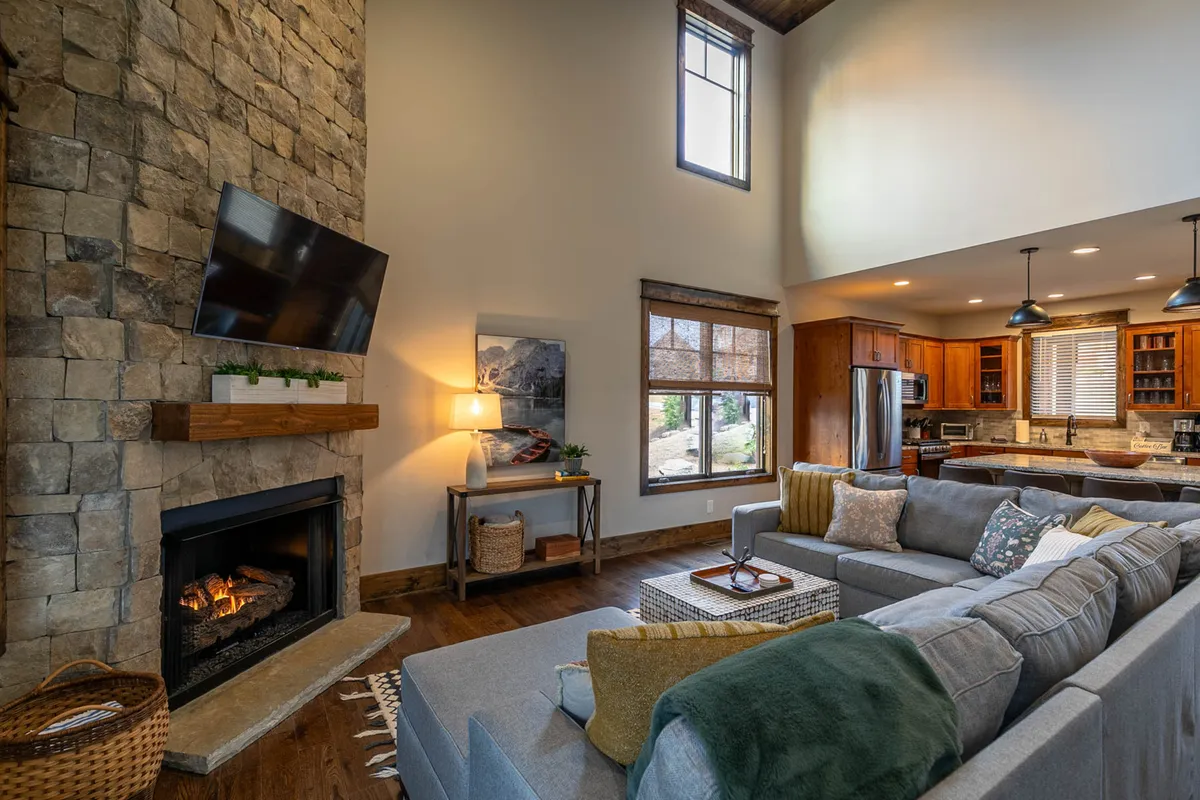 A warm, welcoming family room with a large sectional sofa facing a contemporary stone fireplace, open to the kitchen area in the background. The space is bathed in natural light from a large window, and the setting encourages group conversation or movie nights.